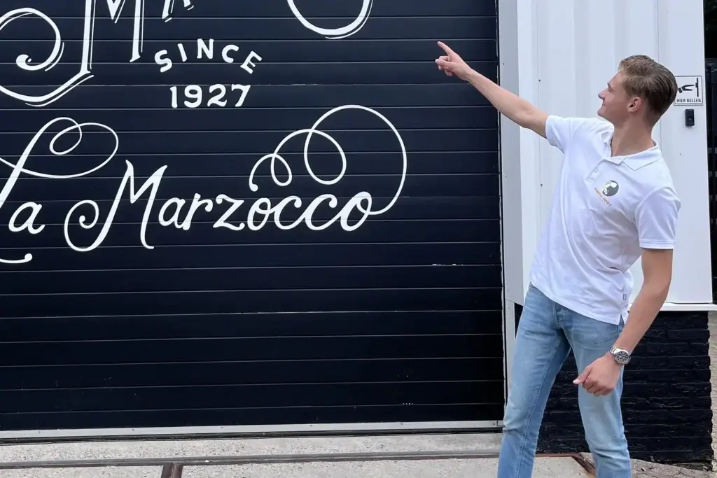 Jordy wijst naar een roldeur met het opschrift van La Marzocco
