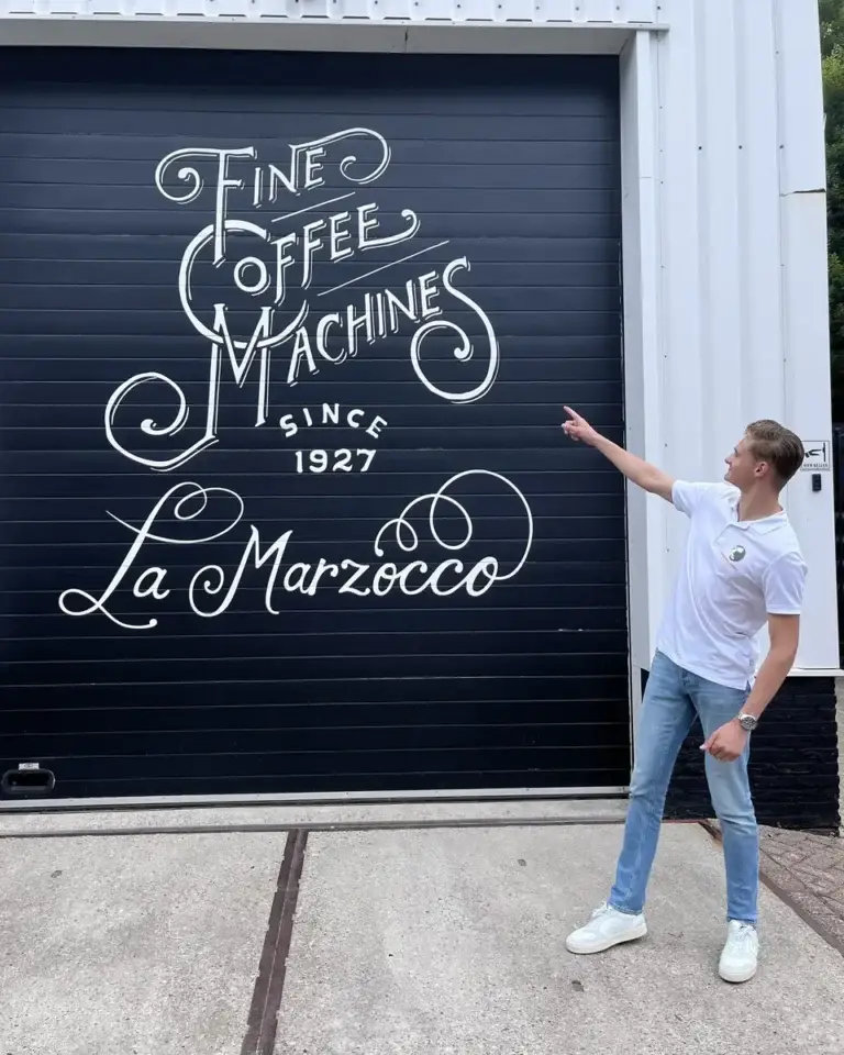Jordy wijst naar een roldeur met het opschrift van La Marzocco
